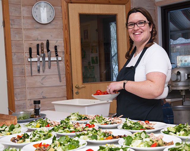 Smilende kvinne i hvit skjorte legger opp mat på tallerken på kjøkkenbenk